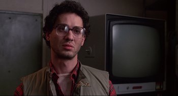 Movie still from “Videodrome” (1983), directed by David Cronenberg – A man wearing glasses and a plaid shirt is standing in front of a television; Close Up shot, Low angle