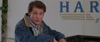 Movie still from “Village of the Damned” (1995), directed by John Carpenter – A man sitting in front of a wall with his arms crossed; Close Up shot, Over the shoulder angle