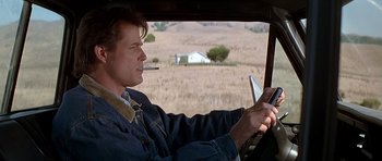 Movie still from “Village of the Damned” (1995), directed by John Carpenter – A man sitting in the driver's seat of a car while smoking a cigarette; Medium shot, Low angle