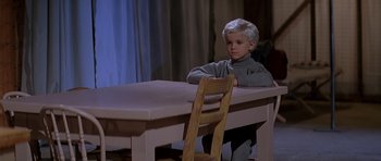 Movie still from “Village of the Damned” (1995), directed by John Carpenter – A boy sitting at a table with a wooden chair in front of him; Medium shot, High angle