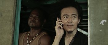 Movie still from “Vinyan” (2008), directed by Fabrice du Welz – A man talking on a cell phone next to another man; Close Up shot, Low angle