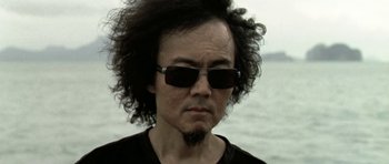 Movie still from “Vinyan” (2008), directed by Fabrice du Welz – A man wearing sunglasses and a black t - shirt looks at the camera; Close Up shot, Low angle