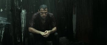 Movie still from “Vinyan” (2008), directed by Fabrice du Welz – A man sitting in the rain with his hands folded on his knees; Medium shot, Low angle