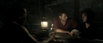 Movie still from “Vinyan” (2008), directed by Fabrice du Welz – A man sitting at a table in a dimly - lit room; Medium shot, Over the shoulder angle