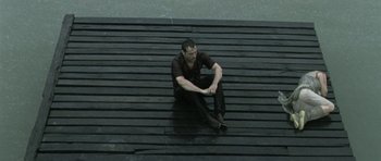 Movie still from “Vinyan” (2008), directed by Fabrice du Welz – A man sitting on a wooden deck on a rainy day; Wide shot, High angle