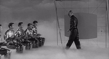 Movie still from “Visit to a Small Planet” (1960), directed by Norman Taurog – A man walking on a stage while two men sit on a bench and one man is sitting on a chair; Wide shot, Low angle