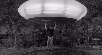 Movie still from “Visit to a Small Planet” (1960), directed by Norman Taurog – A man holding a large metal object above his head; Wide shot, Low angle