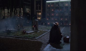 Movie still from “Voice of the Moon” (1990), directed by Richard Stanley – A woman in a black coat and a hat is in front of a grave; Wide shot, High angle