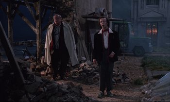 Movie still from “Voice of the Moon” (1990), directed by Richard Stanley – A couple of men standing next to each other on a dirt field; Wide shot, Low angle