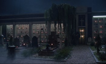 Movie still from “Voice of the Moon” (1990), directed by Richard Stanley – A cemetery with a fountain and a building in the background at night; Wide shot, High angle