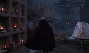 Movie still from “Voice of the Moon” (1990), directed by Richard Stanley – A man wearing a black coat and a hat walking down a street; Wide shot, Low angle