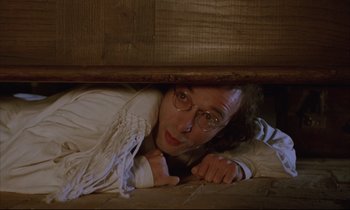 Movie still from “Voice of the Moon” (1990), directed by Richard Stanley – A man with glasses hiding under a bed; Medium shot, Low angle