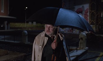 Movie still from “Voice of the Moon” (1990), directed by Richard Stanley – An older man holding an umbrella while standing on a sidewalk; Medium shot, Low angle