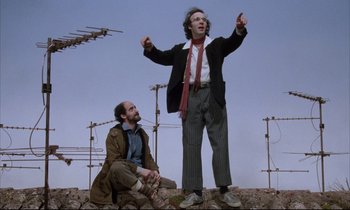 Movie still from “Voice of the Moon” (1990), directed by Richard Stanley – Two men are sitting on the ground and one is holding a microphone; Medium shot, Low angle