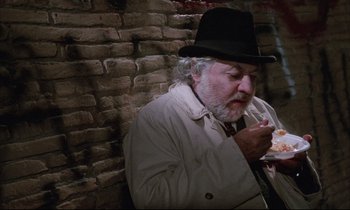 Movie still from “Voice of the Moon” (1990), directed by Richard Stanley – An older man wearing a black hat eating a piece of cake; Medium shot, High angle