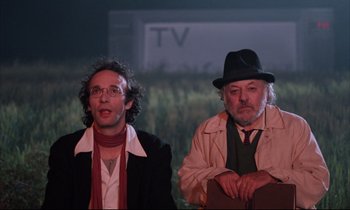 Movie still from “Voice of the Moon” (1990), directed by Richard Stanley – A couple of men sitting next to each other on a stage; Medium shot, Low angle