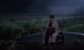 Movie still from “Voice of the Moon” (1990), directed by Richard Stanley – An old man sitting on the side of the road at night; Wide shot, High angle
