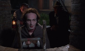 Movie still from “Voice of the Moon” (1990), directed by Richard Stanley – A man in a costume sitting in front of a wooden chair; Medium shot, Over the shoulder angle