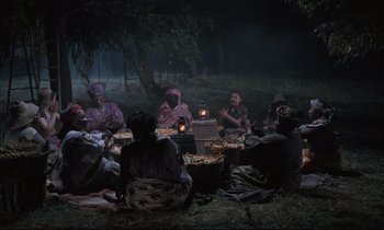 Movie still from “Voice of the Moon” (1990), directed by Richard Stanley – A group of people sitting around a fire pit at night; Wide shot, High angle