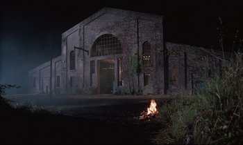 Movie still from “Voice of the Moon” (1990), directed by Richard Stanley – An old building with a fire in front of it at night; Extreme Wide shot, Low angle