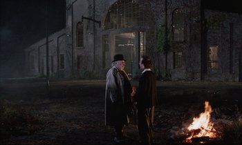 Movie still from “Voice of the Moon” (1990), directed by Richard Stanley – Two men standing in front of a fire at night; Wide shot, Over the shoulder angle