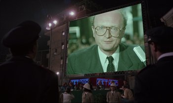 Movie still from “Voice of the Moon” (1990), directed by Richard Stanley – A man in a suit and glasses is on a large screen; Close Up shot, Over the shoulder angle