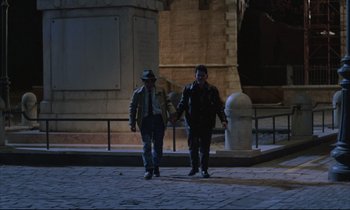 Movie still from “Voice of the Moon” (1990), directed by Richard Stanley – Two men walking on a sidewalk holding hands; Wide shot, High angle