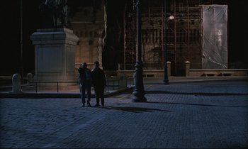 Movie still from “Voice of the Moon” (1990), directed by Richard Stanley – Two people are standing on the sidewalk at night; Extreme Wide shot, High angle