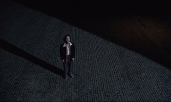 Movie still from “Voice of the Moon” (1990), directed by Richard Stanley – A man standing on a cobblestone street at night; Extreme Wide shot, Overhead angle