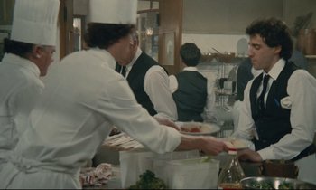 Movie still from “Waiter!” (1983), directed by Claude Sautet – A group of people standing around a table; Medium shot, Over the shoulder angle