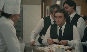 Movie still from “Waiter!” (1983), directed by Claude Sautet – A group of men standing around a table; Medium shot, Over the shoulder angle