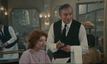 Movie still from “Waiter!” (1983), directed by Claude Sautet – An older man in a vest is writing on a piece of paper while a young woman sits in front of him; Medium shot, Over the shoulder angle