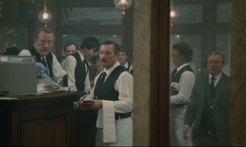 Movie still from “Waiter!” (1983), directed by Claude Sautet – A group of people in a room with a man in a white shirt; Medium shot, Over the shoulder angle