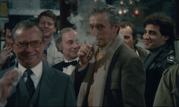 Movie still from “Waiter!” (1983), directed by Claude Sautet – An older man smoking a cigarette in front of a group of people; Medium shot, Over the shoulder angle