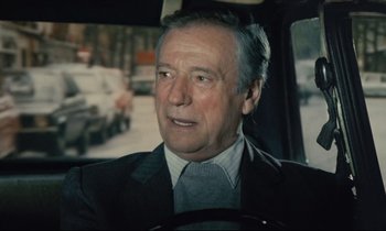 Movie still from “Waiter!” (1983), directed by Claude Sautet – An older man wearing a suit and tie driving a car; Close Up shot, Over the shoulder angle