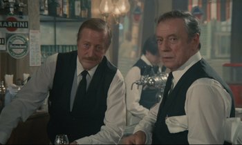 Movie still from “Waiter!” (1983), directed by Claude Sautet – A couple of men sitting next to each other in a room; Medium shot, Over the shoulder angle