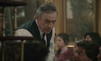 Movie still from “Waiter!” (1983), directed by Claude Sautet – An older man in a vest and tie is sitting at a table; Medium shot, Over the shoulder angle