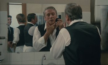 Movie still from “Waiter!” (1983), directed by Claude Sautet – An older man is looking in the bathroom mirror while putting on his tie; Medium shot, Over the shoulder angle
