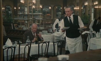 Movie still from “Waiter!” (1983), directed by Claude Sautet – A man in a suit and tie standing at a table with a tray of food in front of him; Wide shot, High angle