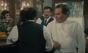 Movie still from “Waiter!” (1983), directed by Claude Sautet – A man in a white shirt and a man in a black vest shaking hands in front of other men; Medium shot, Over the shoulder angle