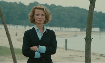 Movie still from “Waiter!” (1983), directed by Claude Sautet – A woman standing in front of a body of water with her arms crossed; Medium shot, Low angle