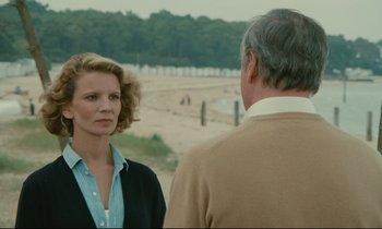Movie still from “Waiter!” (1983), directed by Claude Sautet – A woman standing next to an older man on the beach; Medium shot, Over the shoulder angle