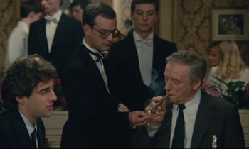 Movie still from “Waiter!” (1983), directed by Claude Sautet – A group of men standing around each other smoking; Medium shot, Over the shoulder angle