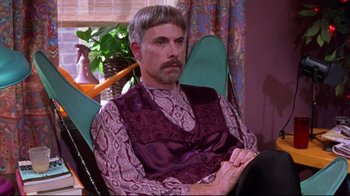 Movie still from “Waiting for Guffman” (1996), directed by Christopher Guest – A man sitting in a chair wearing a purple vest; Medium shot, Over the shoulder angle