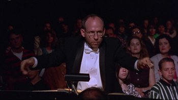 Movie still from “Waiting for Guffman” (1996), directed by Christopher Guest – A man wearing a tuxedo and bow tie is holding a conductor's baton in front of an audience; Medium shot, Low angle