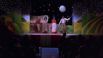 Movie still from “Waiting for Guffman” (1996), directed by Christopher Guest – A group of people standing on a stage in front of a screen; Wide shot, High angle