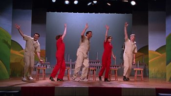 Movie still from “Waiting for Guffman” (1996), directed by Christopher Guest – A group of people on a stage with one person holding his hands up; Wide shot, High angle