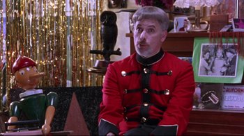 Movie still from “Waiting for Guffman” (1996), directed by Christopher Guest – A man in a red and black uniform sitting on a chair; Medium shot, Over the shoulder angle
