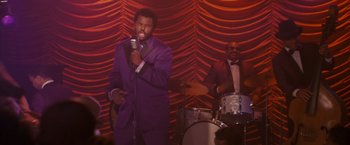 Movie still from “Walk Hard: The Dewey Cox Story” (2007), directed by Jake Kasdan – A man in a purple suit singing into a microphone; Medium shot, Over the shoulder angle