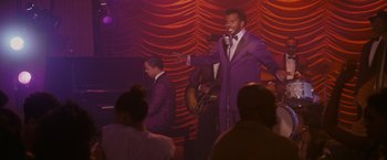 Movie still from “Walk Hard: The Dewey Cox Story” (2007), directed by Jake Kasdan – A man in a purple suit singing into a microphone on a stage; Medium shot, Over the shoulder angle
